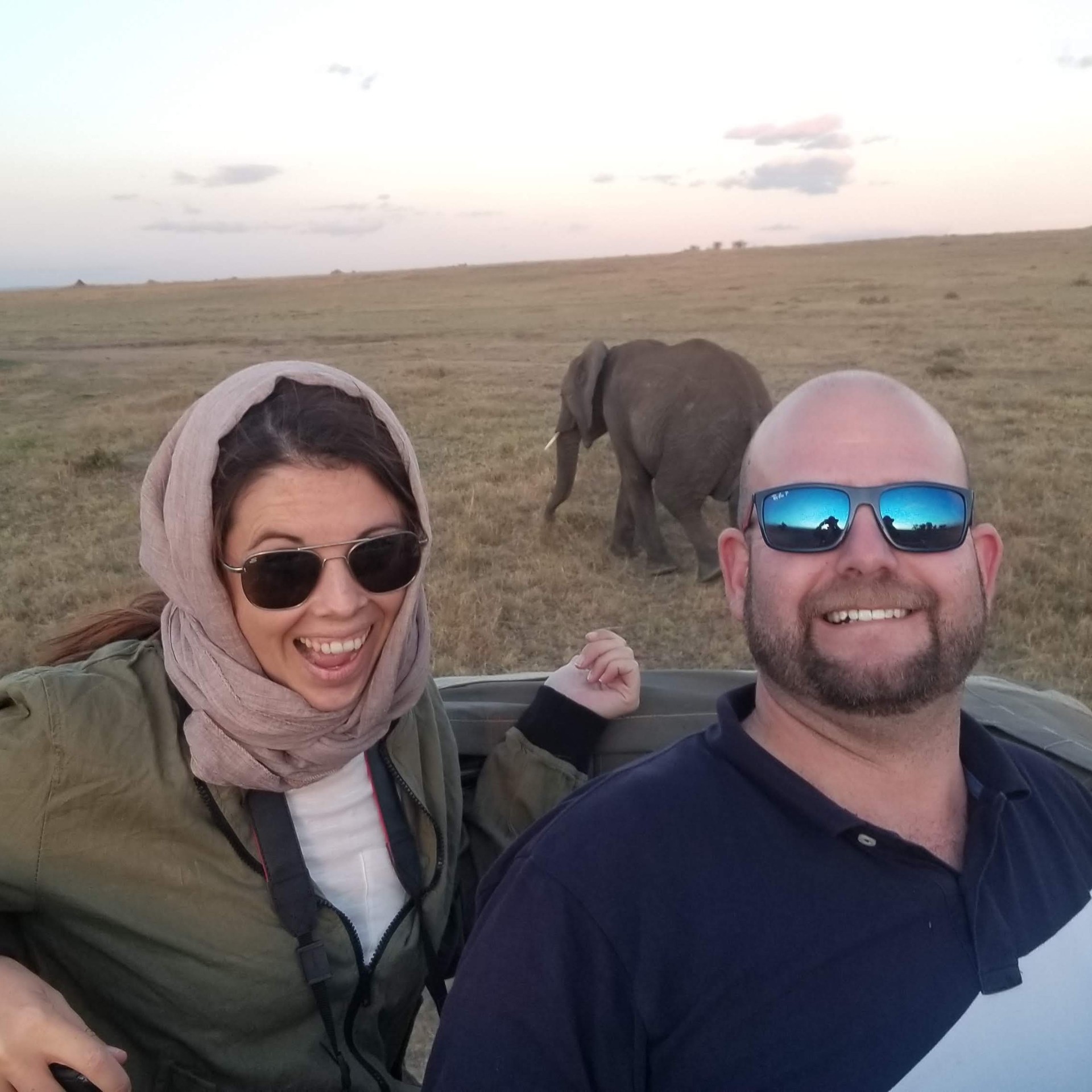 Couple smiling in a safari vehicle with an elephant walking behind them
