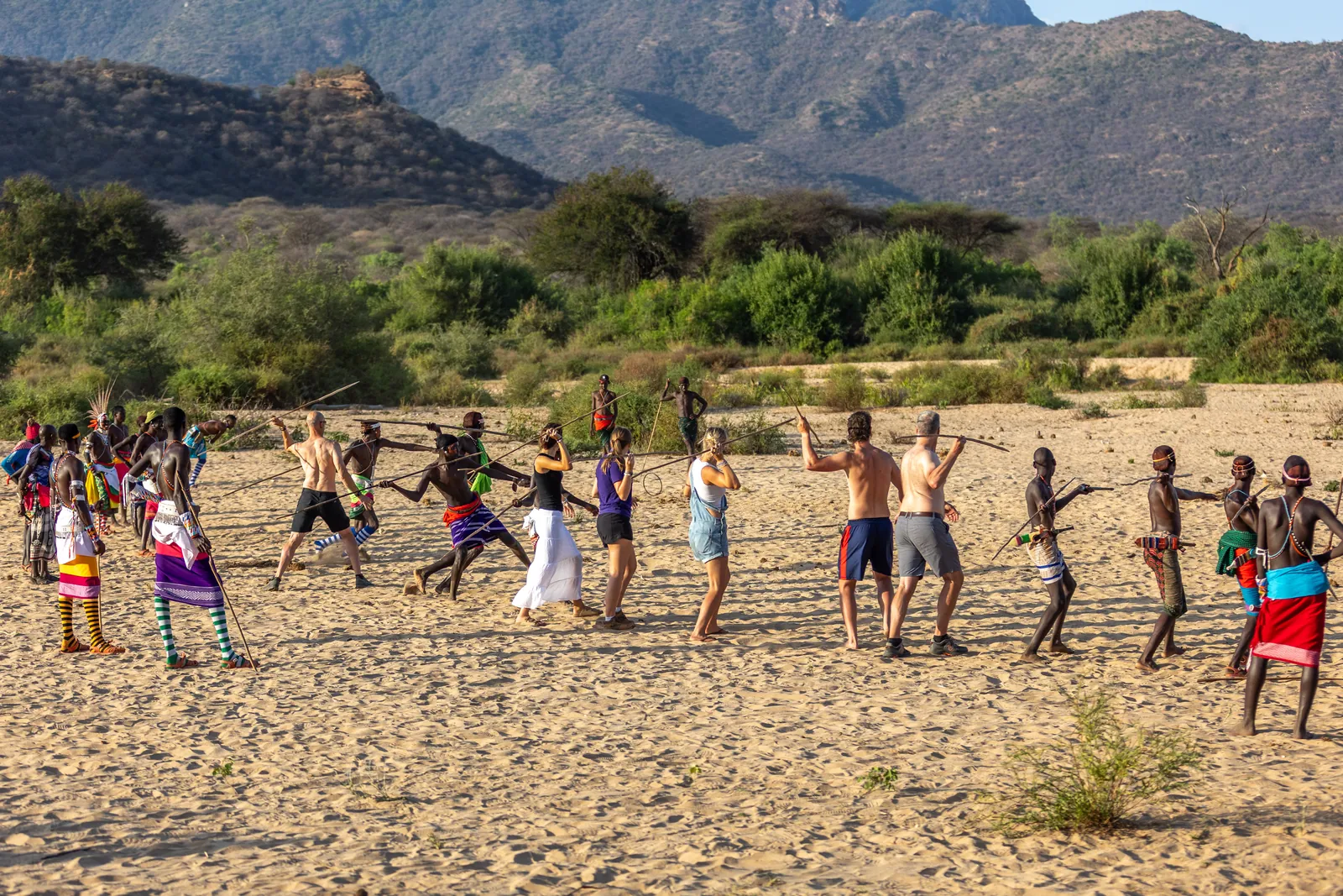 Travelers learning to throw spears with Samburu warriors on a sandy riverbed