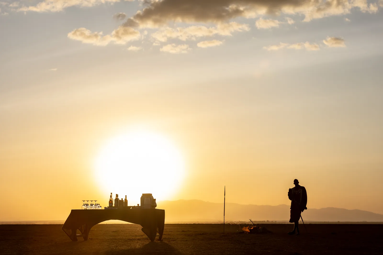 Maasai host tending a fire beside a sundowner bar at sunset